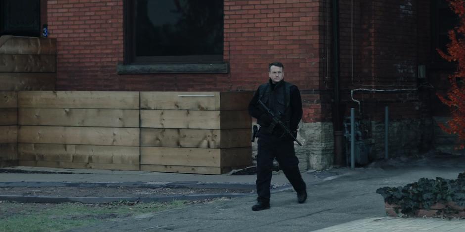 A guard walks down a driveway in front of a house carrying a large gun and looking down the street.