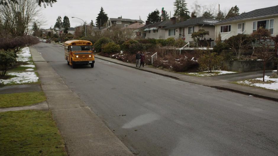 The school bus drives down the street towards the bus stop.