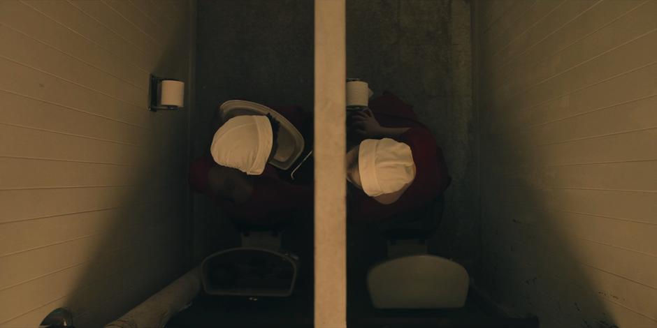 June and Moira sit in adjacent bathroom stalls while Moira carves on the wall and June advises caution.