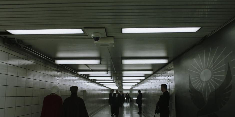 June and Moira walk down a hallway lined with guards past a few other commuters.