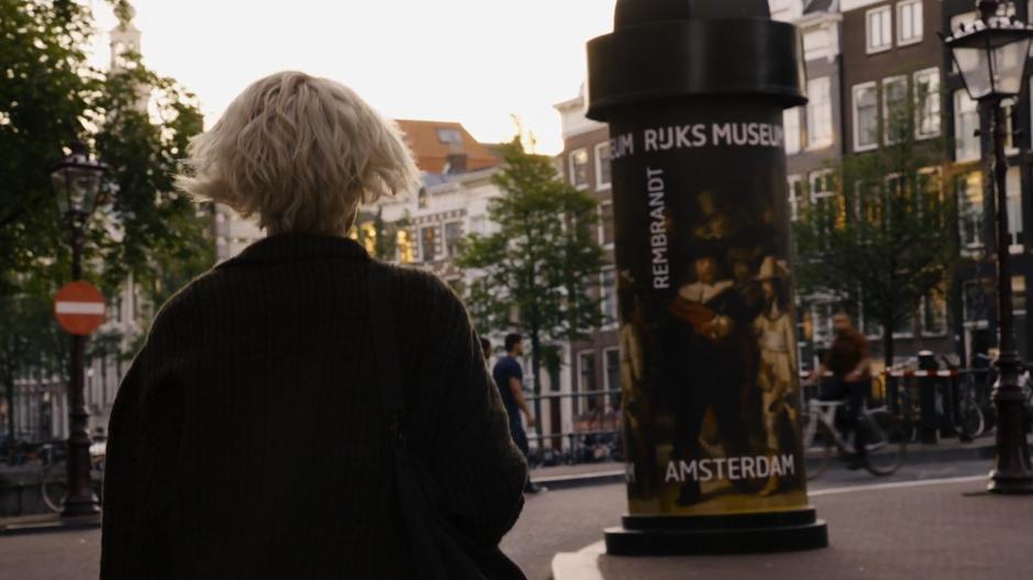 Riley walks down the street past a sign advertising for the Rijksmuseum.