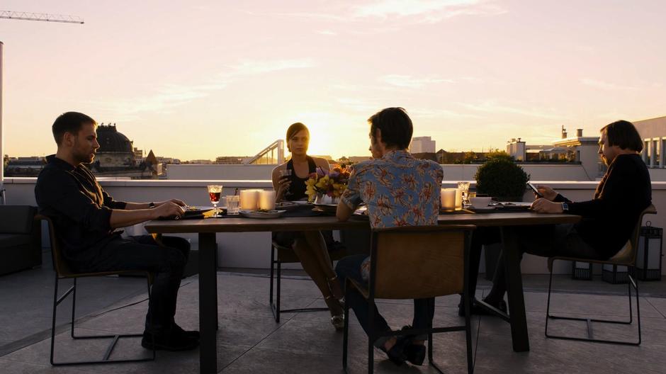 Wolfgang, Lila, Felix, and Sebastian sit at the table talking in front of the sunset after finishing their meal.