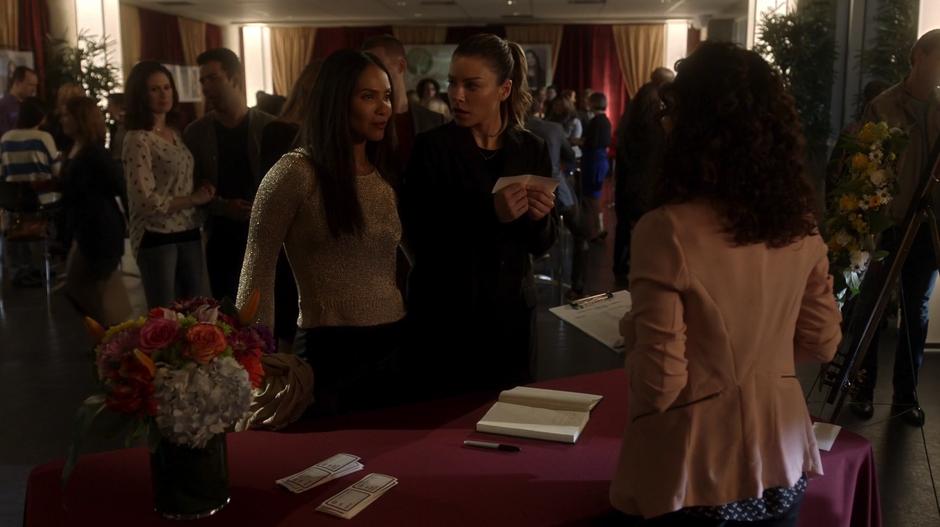 Maze introduces herself as Chloe's wife at the check-in desk while Chloe looks at her with surprise.