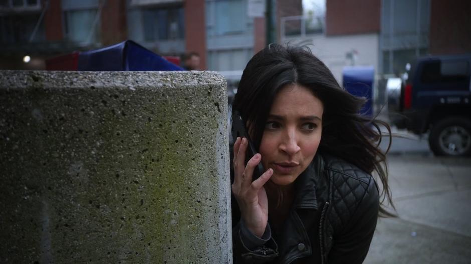 Maggie talks to Alex on the phone while taking cover behind a concrete wall.