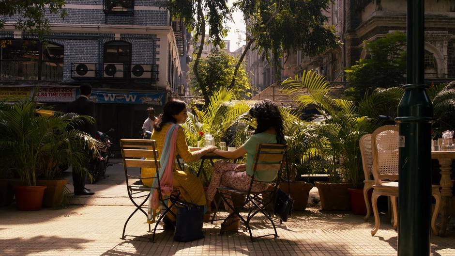 Priya holds Kala's hand while they sit at the cafe table.