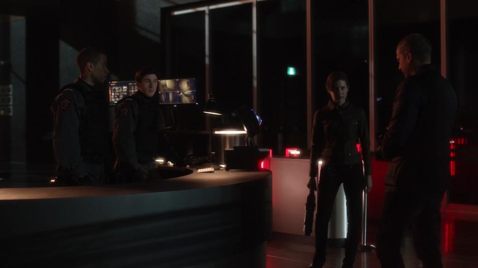 Two guards stand at the desk as Barry using the form of Lyla shows them Snart.