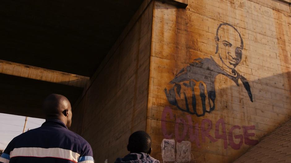 Capheus and Jala look up at graffiti of Capheus with the word Courage written below it.