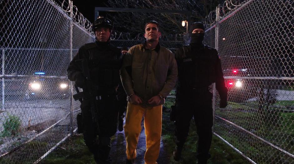 Adrian Chase is escorted by two A.R.G.U.S. guards away from the ground transport.