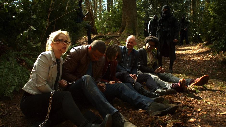 The five captured members of Team Arrow look around the woods while they are chained together.