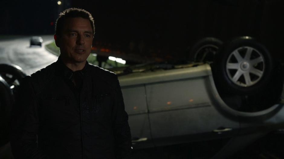 Merlyn talks to Oliver while standing in front of the crashed car.