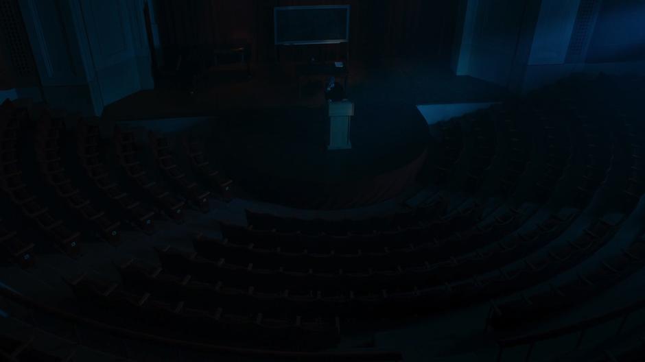 The Doctor stands at the lectern in front of the empty and dark lecture hall.