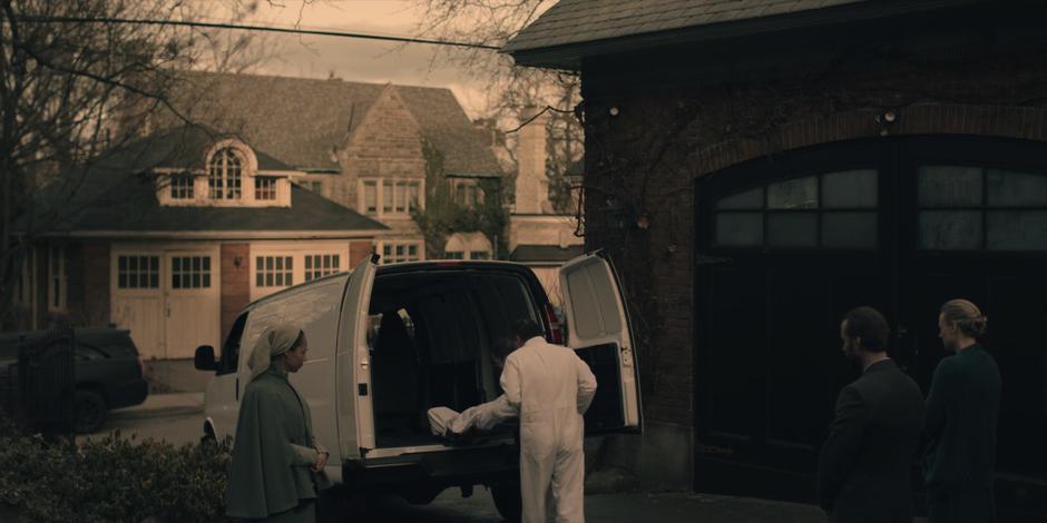 Two men load the former Offred's body into a van in front of the Martha, Fred, & Serena.