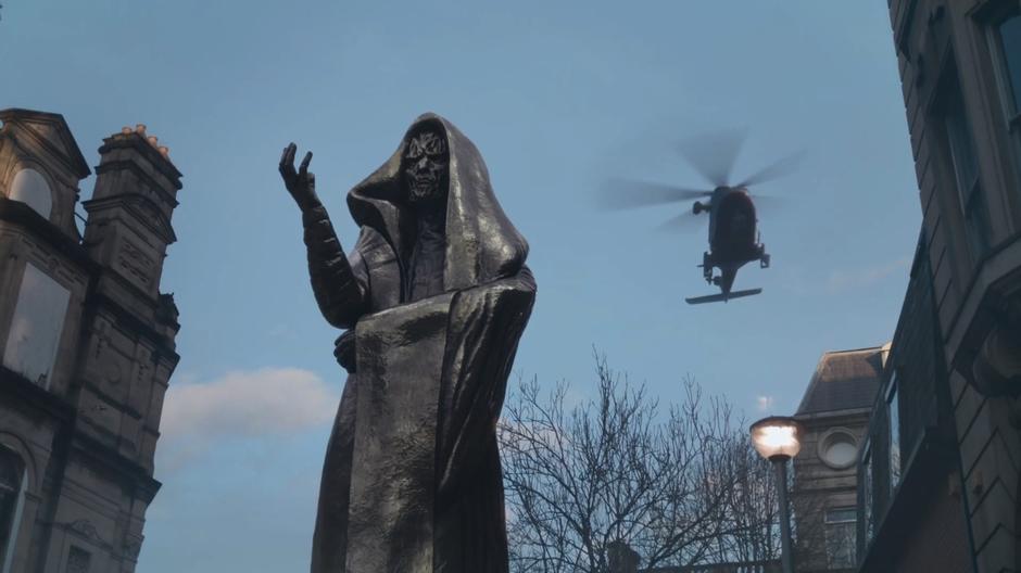 A helicopter flies through the sky behind a monk statue.