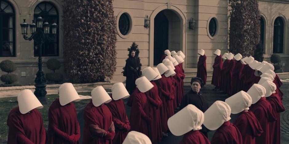 Aunt Lydian looks down the line of handmaids assembled out front to where Janine is getting into the van.