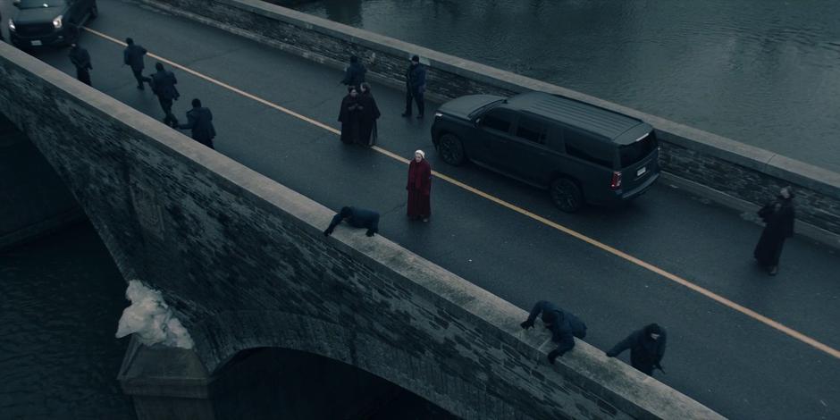 Offred stands stunned in the middle of the bridge while the guards point to the water where Janine fell.