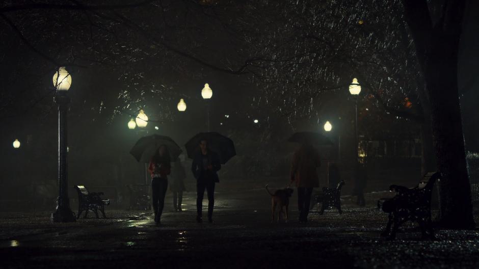 Izzy and Simon stroll through the park in the rain at night with their umbrellas.
