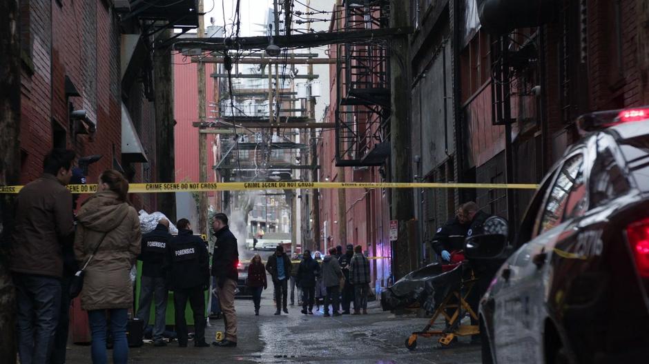 Liv, Ravi, and Clive walk down the alley to where the forensics team is standing around the body.