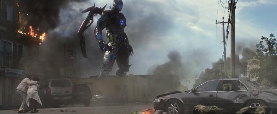 The Megazord stands in the distance while two people walk down the smoky street.