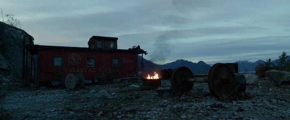 Zack lies atop an abandoned caboose high above the mine while a fire burns in a steel drum in the foreground.