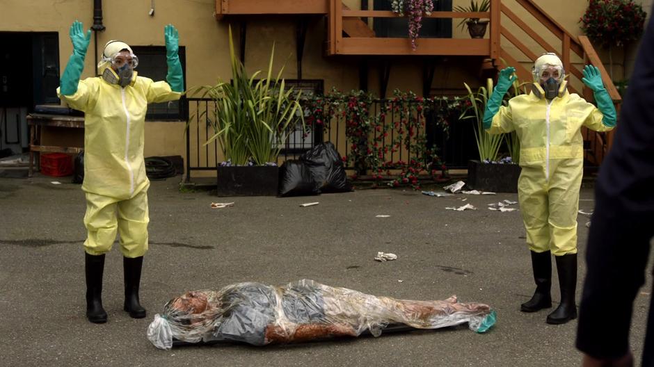 The two cleaning women hold up their hands while standing on either side of a body wrapped in plastic.
