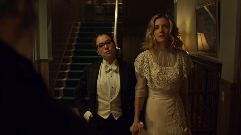 Cosima in a tux and Delphine in a dress stand in the entry hall holding hands.