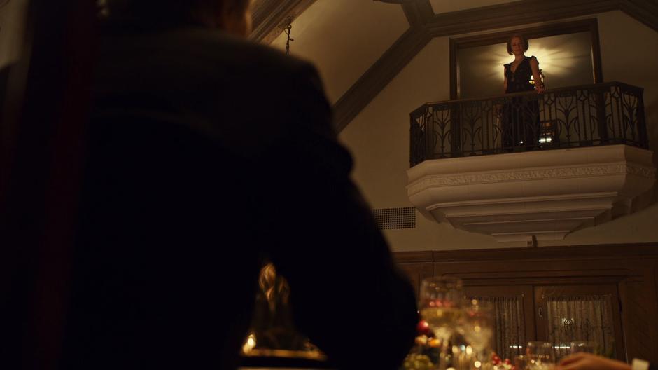 Rachel in a black dress stands on the balcony overlooking the dining room.
