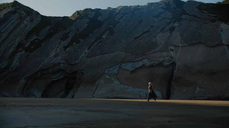 Daenerys strides across the beach in front of the cliffs.