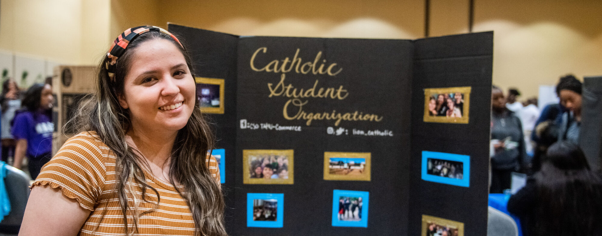 Student smiling with behind a poster with photos of the Catholic Student Organization.