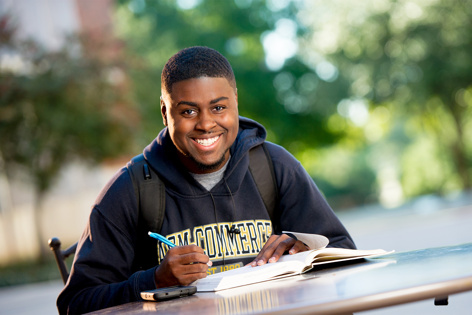 Student smiling siting at a outside table with book in front of him.