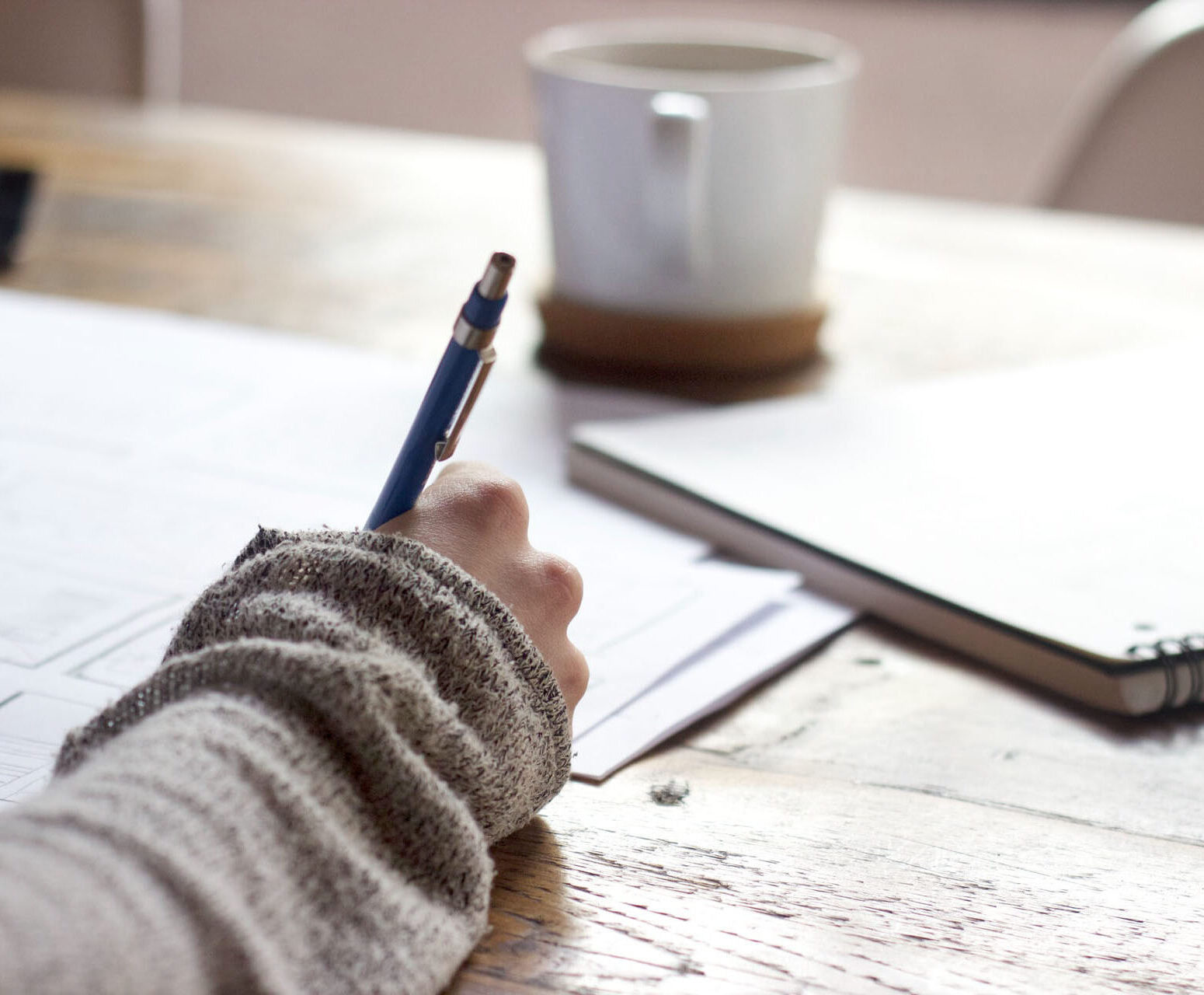 Student writing on the table with mug and and notebook in front.