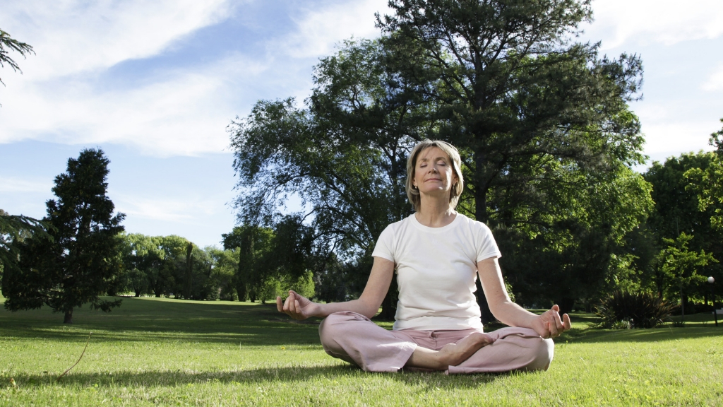 Woman sitting on the green, grassy ground, meditating in a park with trees in the background.