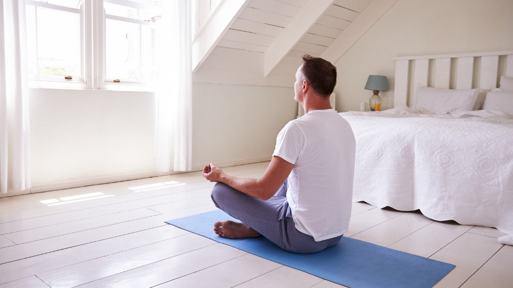 Man sitting on the floor of a serene bedroom with natural light, plants, and minimal clutter.