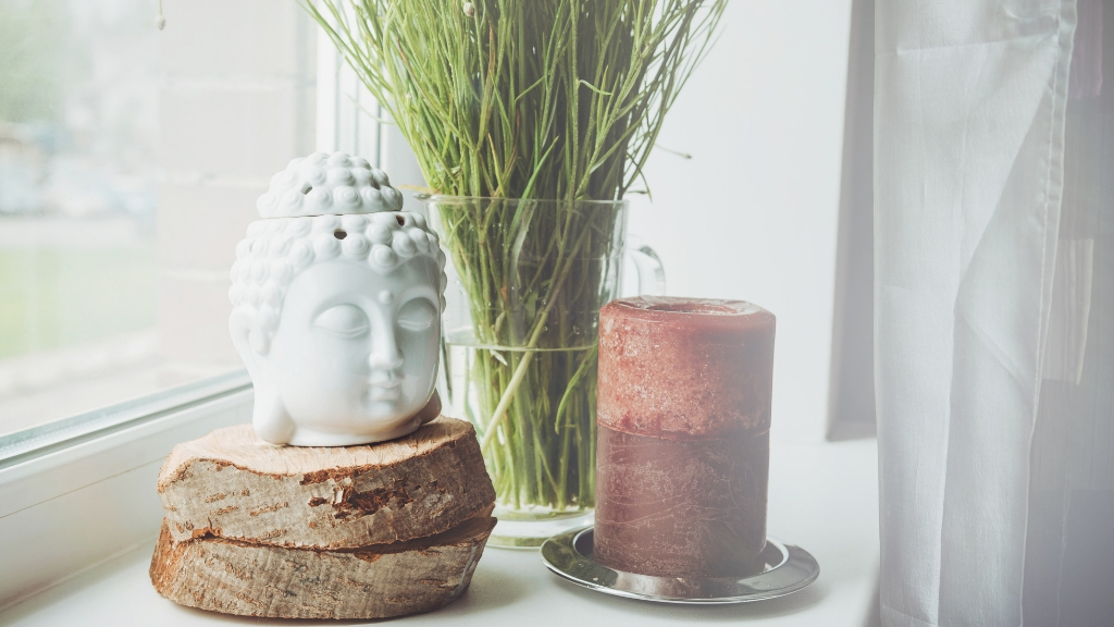 A Buddha head statue, a plant and a large red candle by the window.
