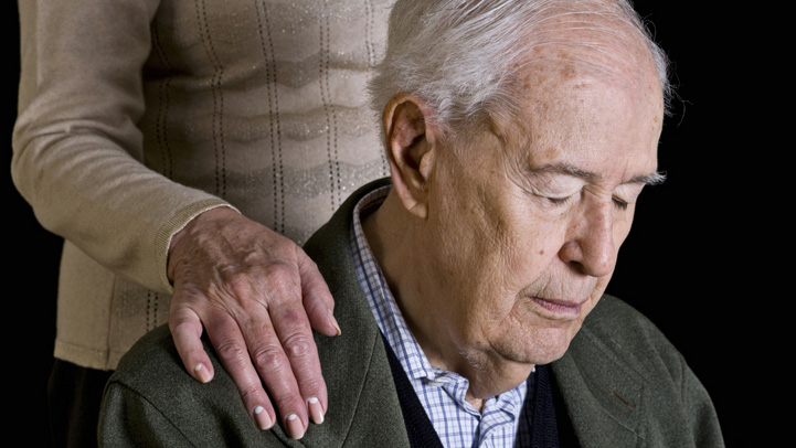 A woman's hand over the shoulder of her husband who is truggling with Huntington's disease