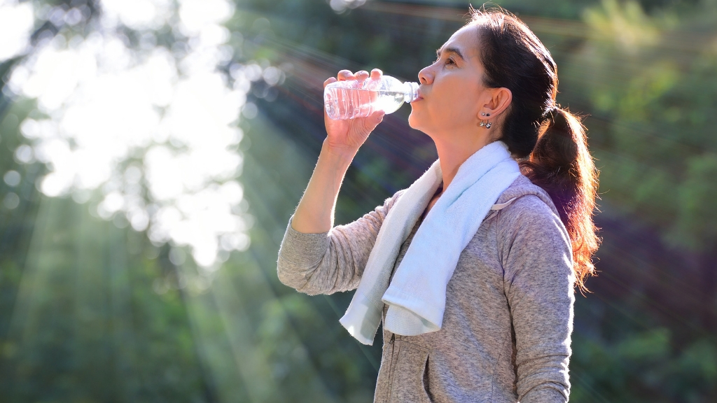 A woman with a towel on her shoulder drinking water from a plastic bottle after exercise.
