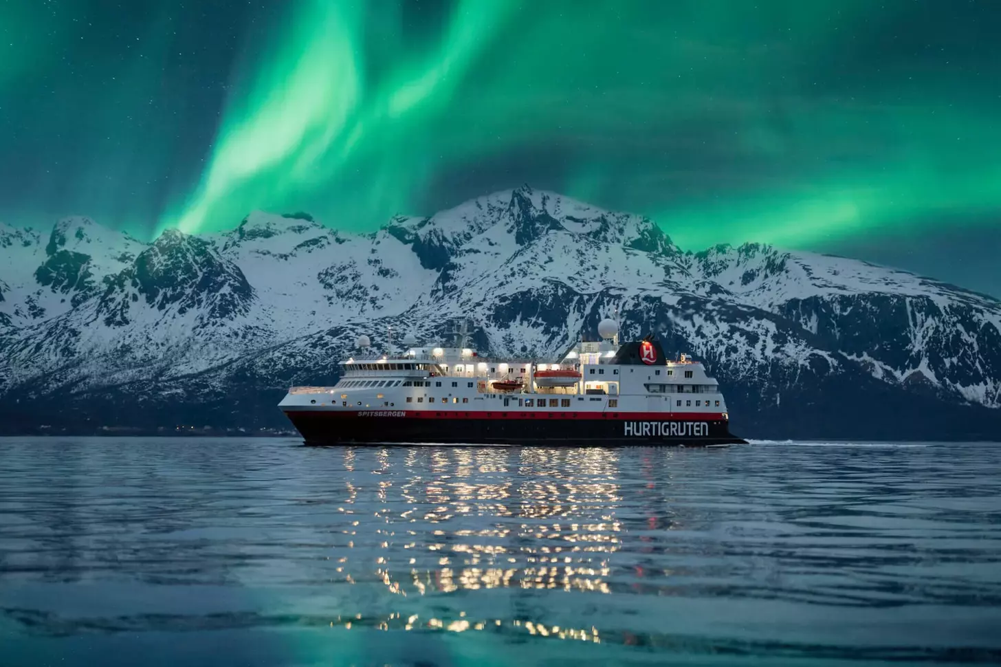 A Hurtigruten cruise ship sailing through Norway with snow covered mountains in the background and green lights across the sky from the stunning Northern Lights