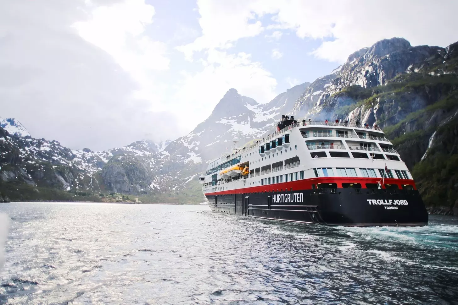 A Hurtigruten ship sailing in wintery conditions with cloud filled skies and snow capped mountains