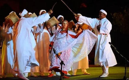 A group of Amazigh dancers performing a traditional folk dance in a lively, rhythmic manner. The dancers move with graceful, fluid motions, their colorful, ornate costumes swirling as they twirl and leap. The background features a rugged, desert-like landscape with towering, ochre-colored cliffs and a vibrant, cloudless sky. Warm, golden lighting illuminates the scene, casting soft shadows and highlighting the energy and dynamism of the performance. The overall mood is one of cultural celebration, authenticity, and deep connection to ancestral traditions.