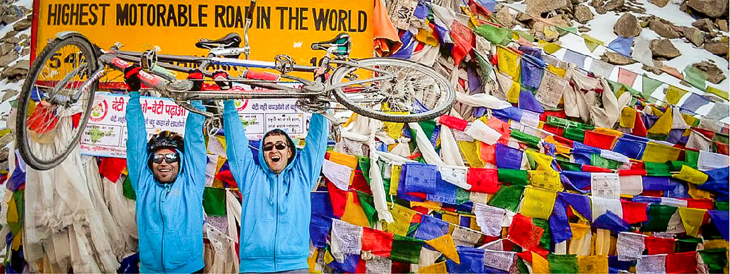 Two riders in blue jackets lift a tandem bicycle over their heads beside a sign reading Highest Motorable Road in the World, surrounded by prayer flags.
