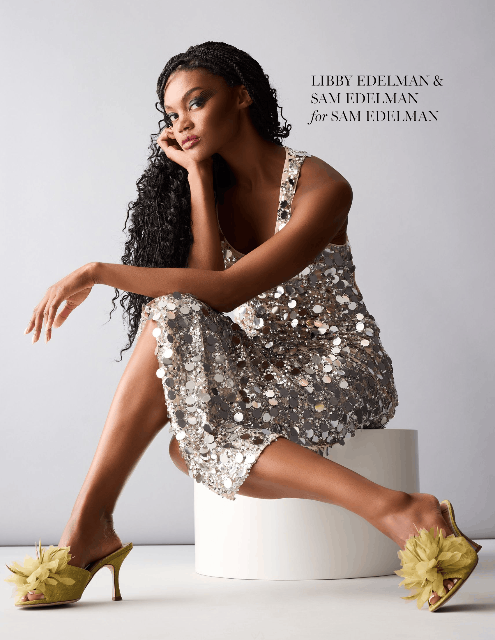 Model Ashley Karah sits on a white cylindrical stool, wearing a sparkling silver sequin dress and yellow floral high-heeled sandals. She poses with one arm resting across her knee against a light gray studio background. Text in the upper right reads, “LIBBY EDELMAN & SAM EDELMAN for SAM EDELMAN.”