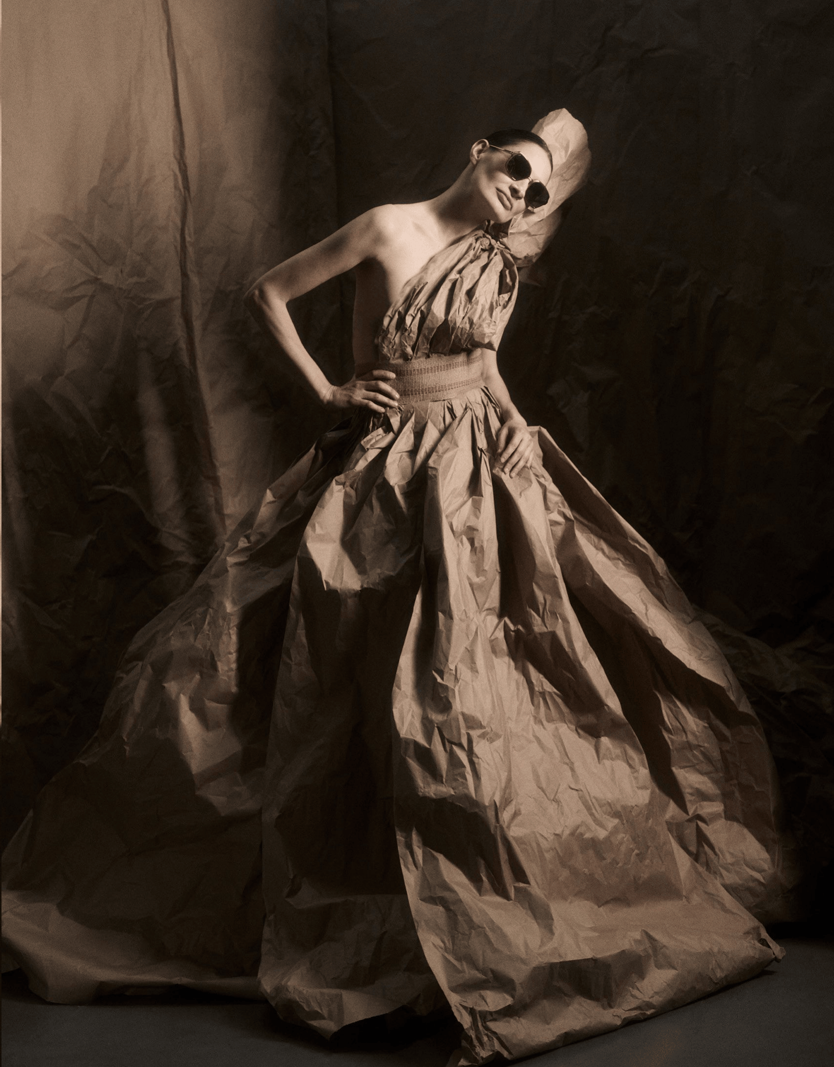 Full-length studio portrait of model Malain Giura wearing narrow sunglasses by Leisure Society and a dramatic voluminous gown made of textured paper material, posed with one hand on her hip