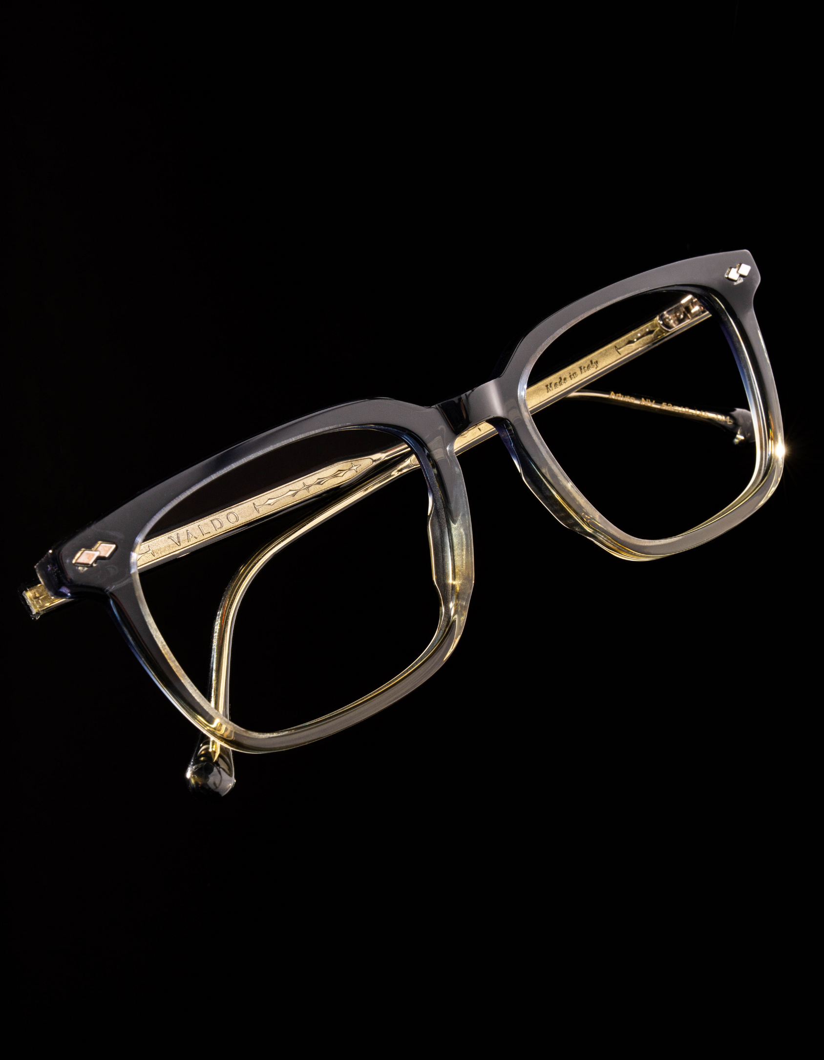 Square translucent gray eyeglasses from Valdo with subtle gradient acetate, gold-tone metal temples, and small diamond-shaped rivet details at the corners, shown against a black background