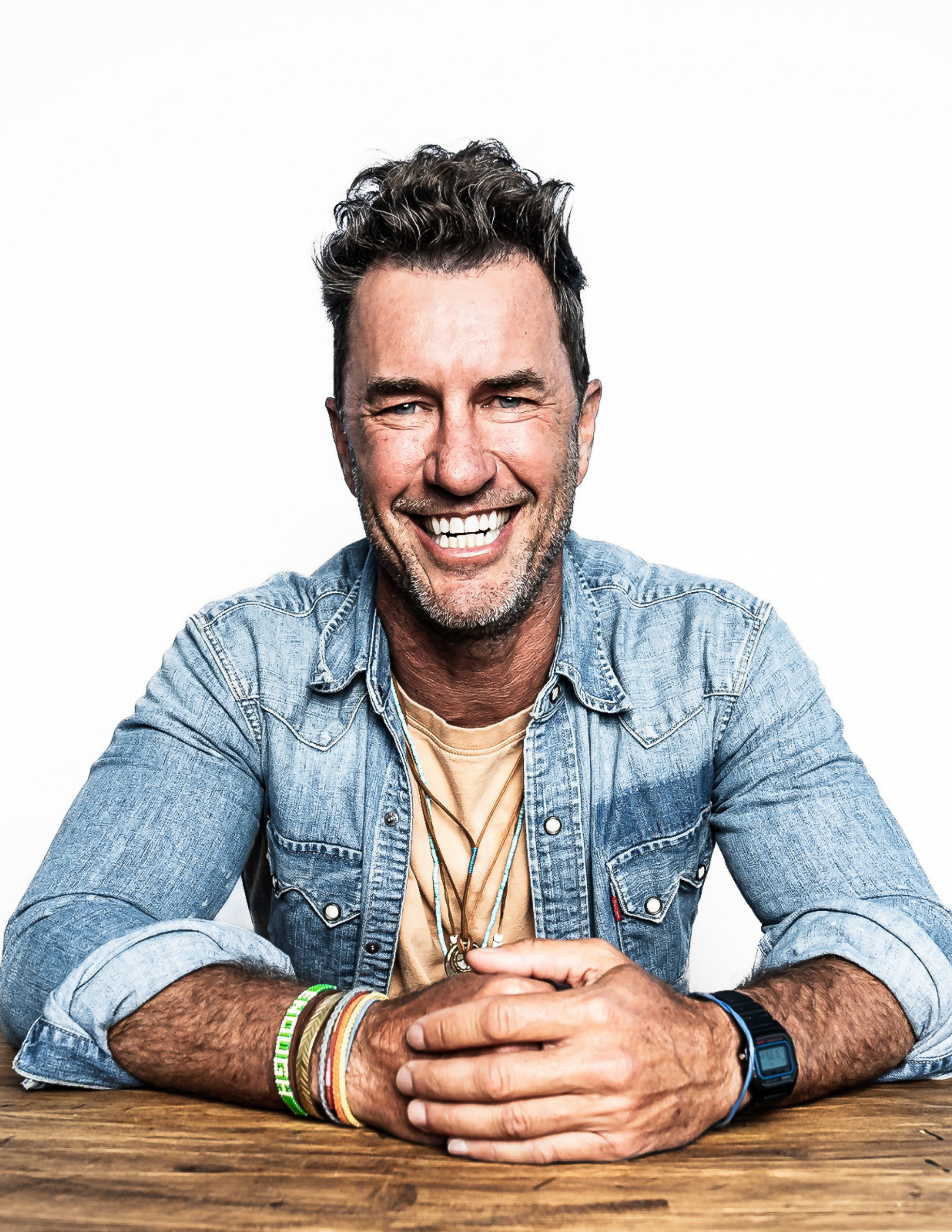 Portrait of Blake Mycoskie seated at a table wearing a denim shirt and stacked We Are Enough bracelets.