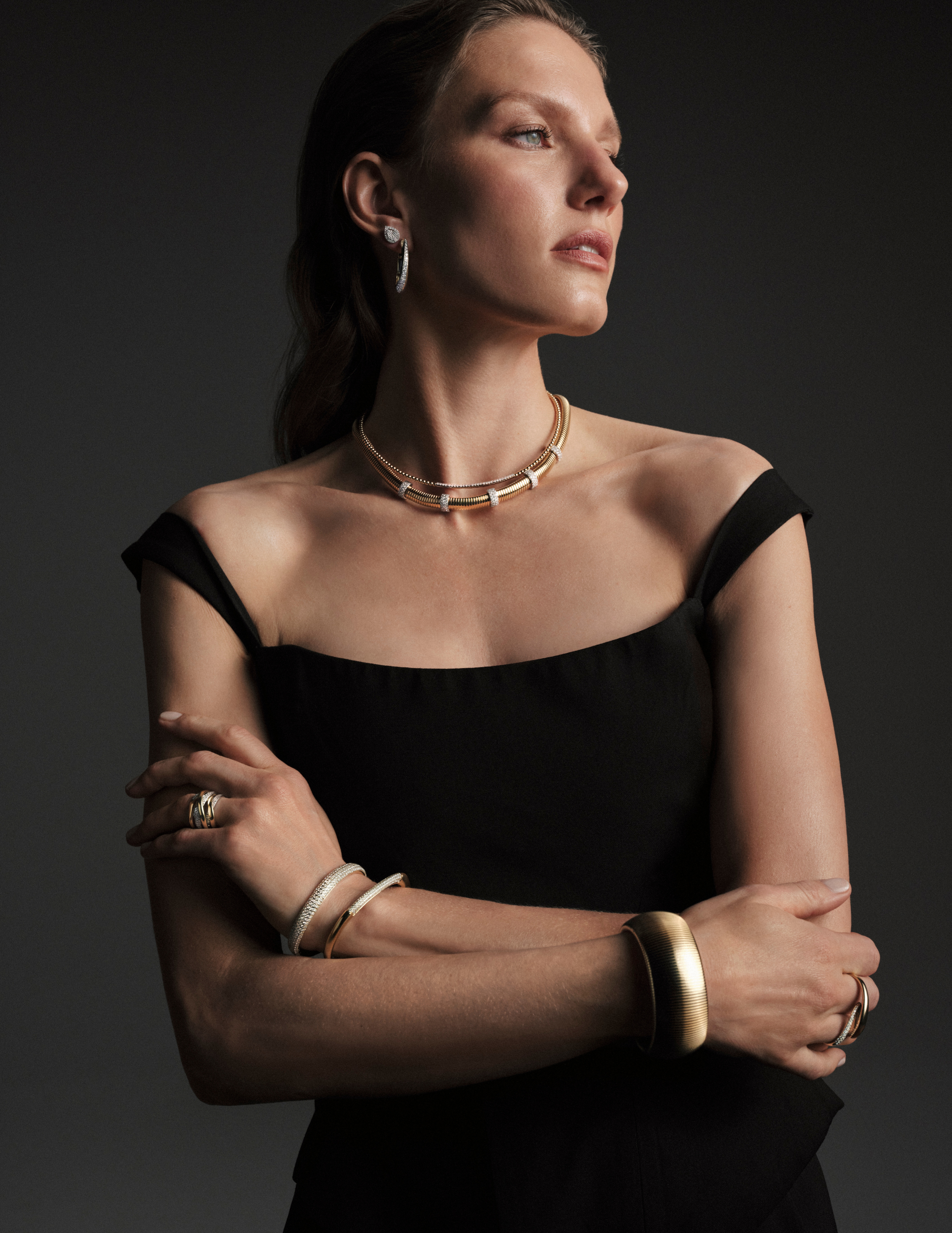 Model wearing an off-the-shoulder black dress styled with layered gold necklaces and bracelets by Simon G. Jewelry