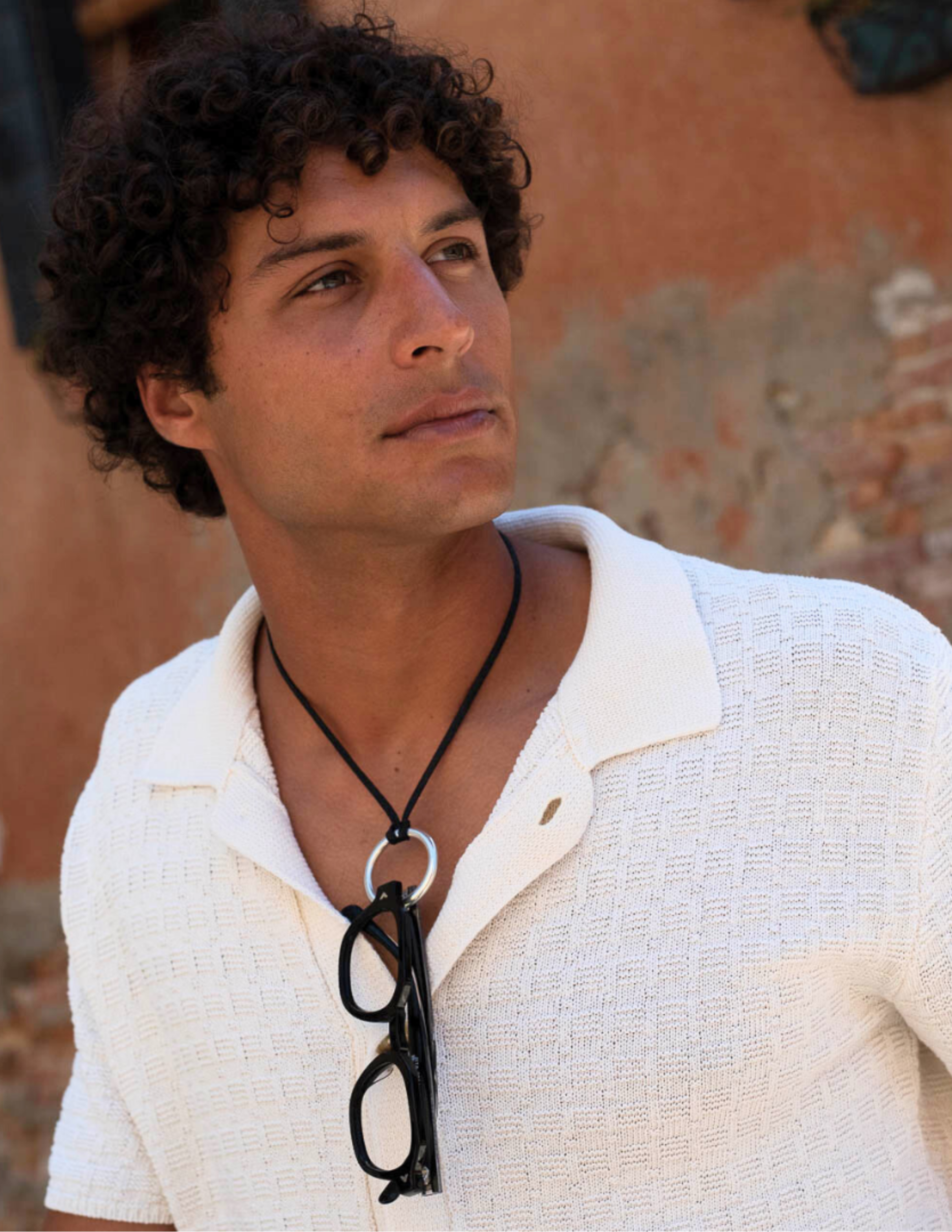 Man wearing a white textured polo shirt with COTI Vision reading glasses suspended from a black neck cord necklace. The portrait highlights the brand's signature blend of eyewear functionality and contemporary style