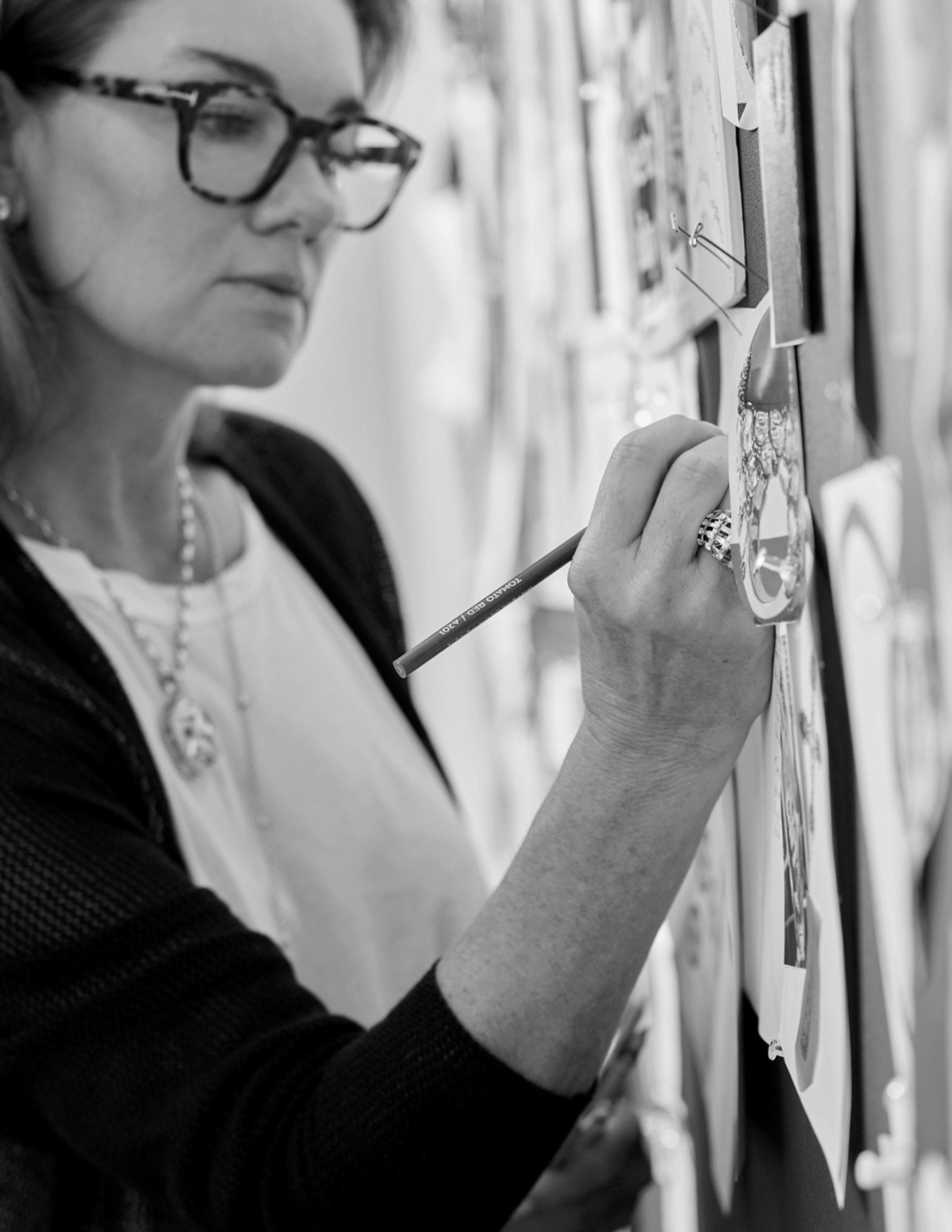 Natalie Katz O’Brien reviews jewelry sketches and inspiration materials pinned to a design board. In this black-and-white image, she is shown working in her studio, illustrating her creative process behind the Natalie O. collection.