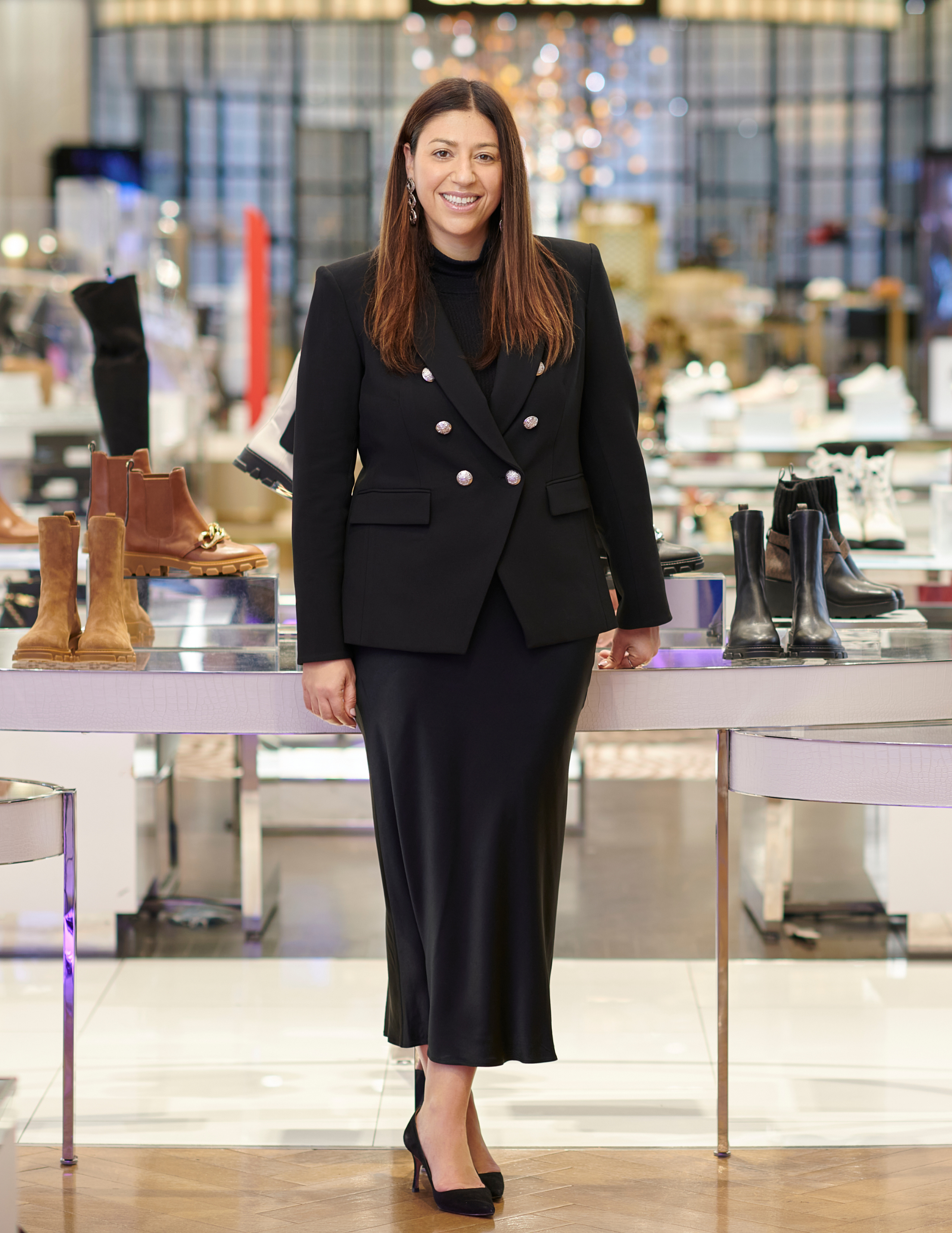 Nata Dvir stands inside a Macy’s store wearing a black blazer and midi skirt, smiling in front of shoe displays in a polished retail environment