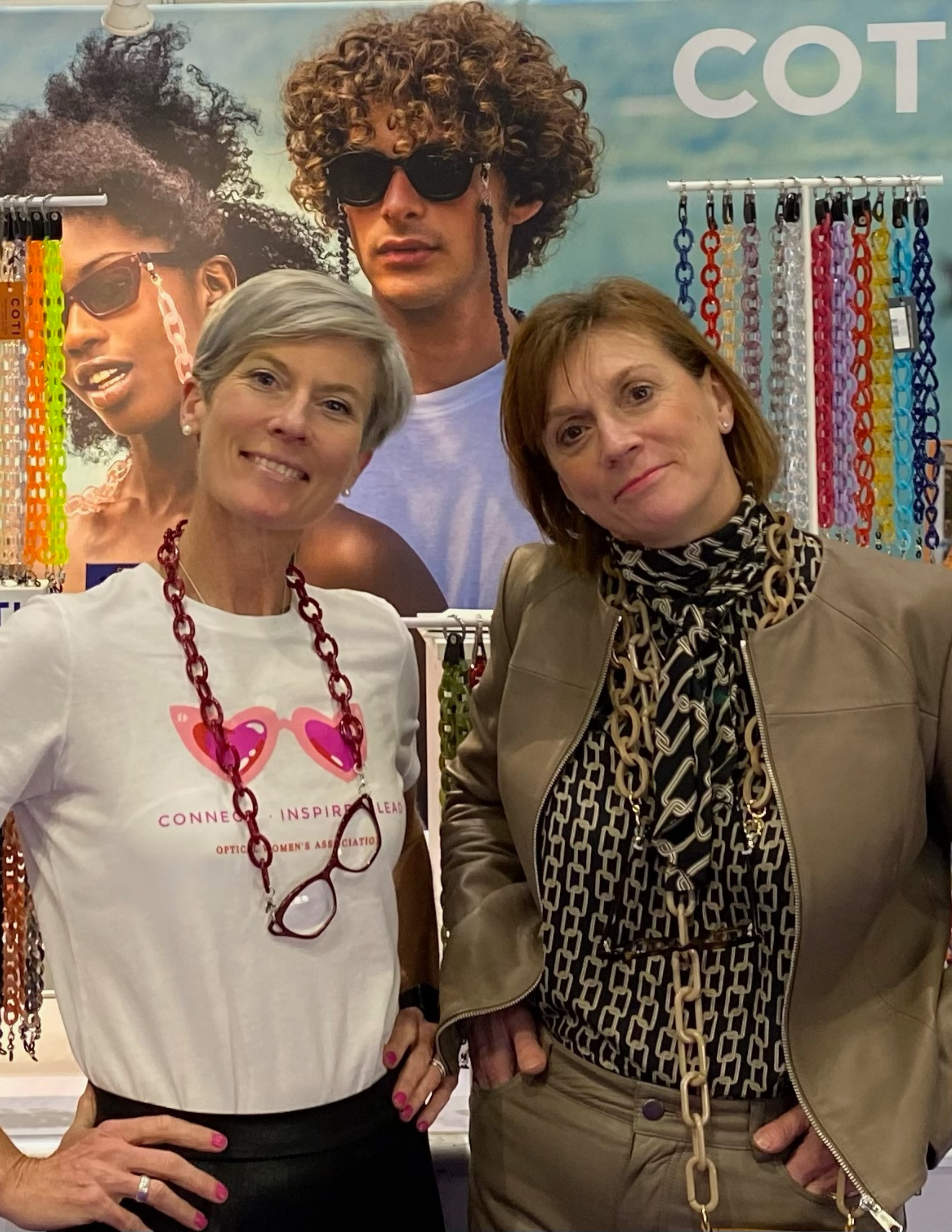 Nancy Gries and Julie Hawkins posing at the COTI Vision trade show booth, wearing the brand's eyewear accessories and reading glass necklaces. The display features colorful eyewear chains and lifestyle imagery that reflect COTI Vision's fashion-focused approach to optical accessories.