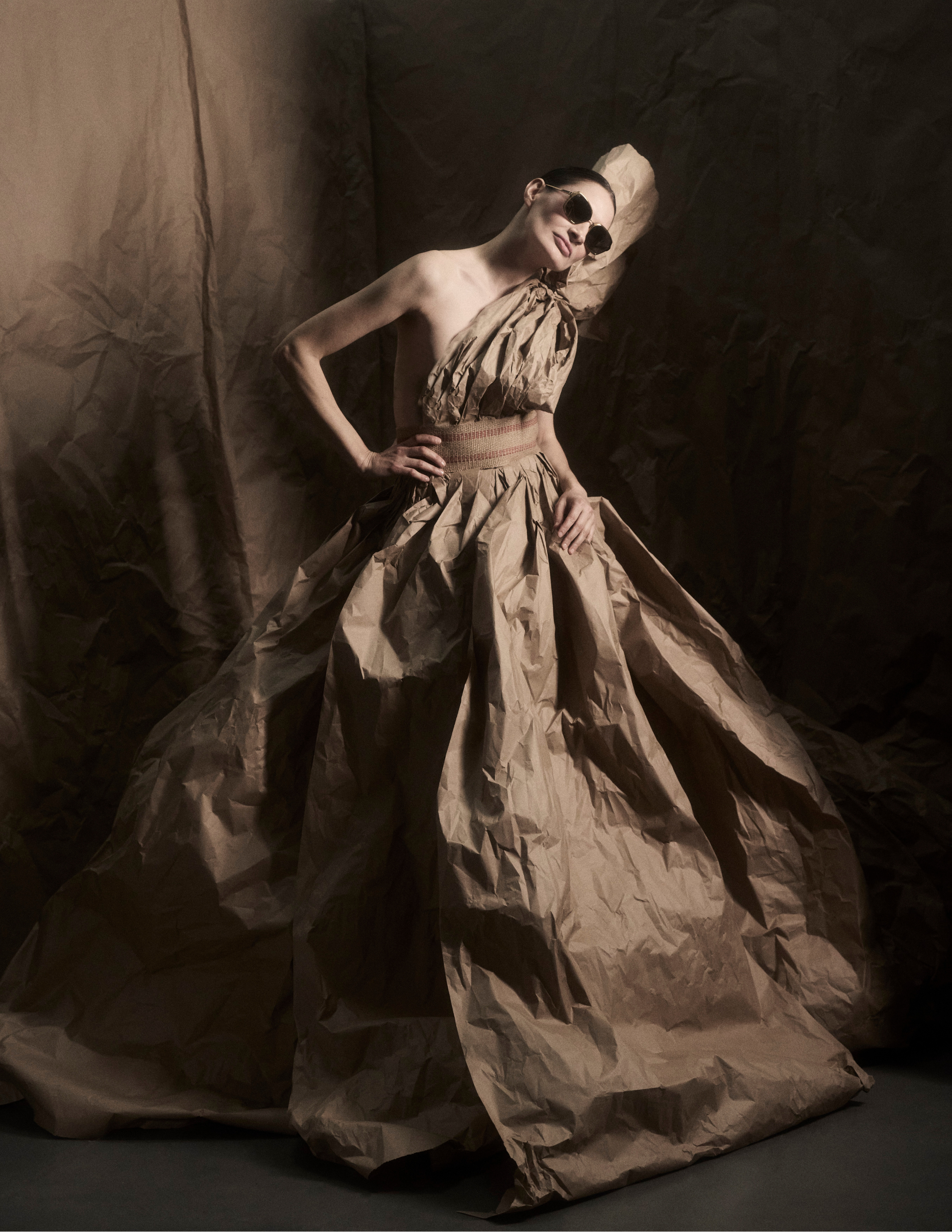 Editorial fashion image featuring a model in a sculptural couture gown crafted from brown paper materials against a dramatic neutral backdrop