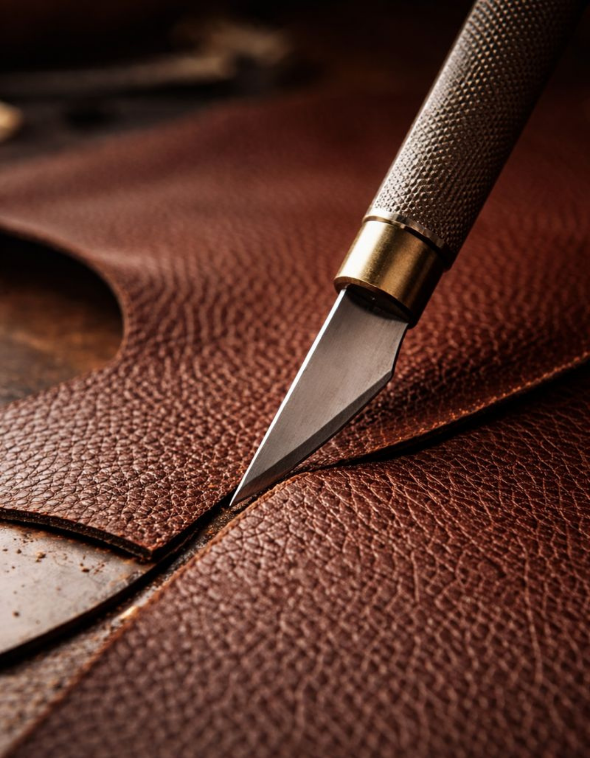 Close-up of a precision craft knife cutting textured brown leather, representing handbag craftsmanship and material design